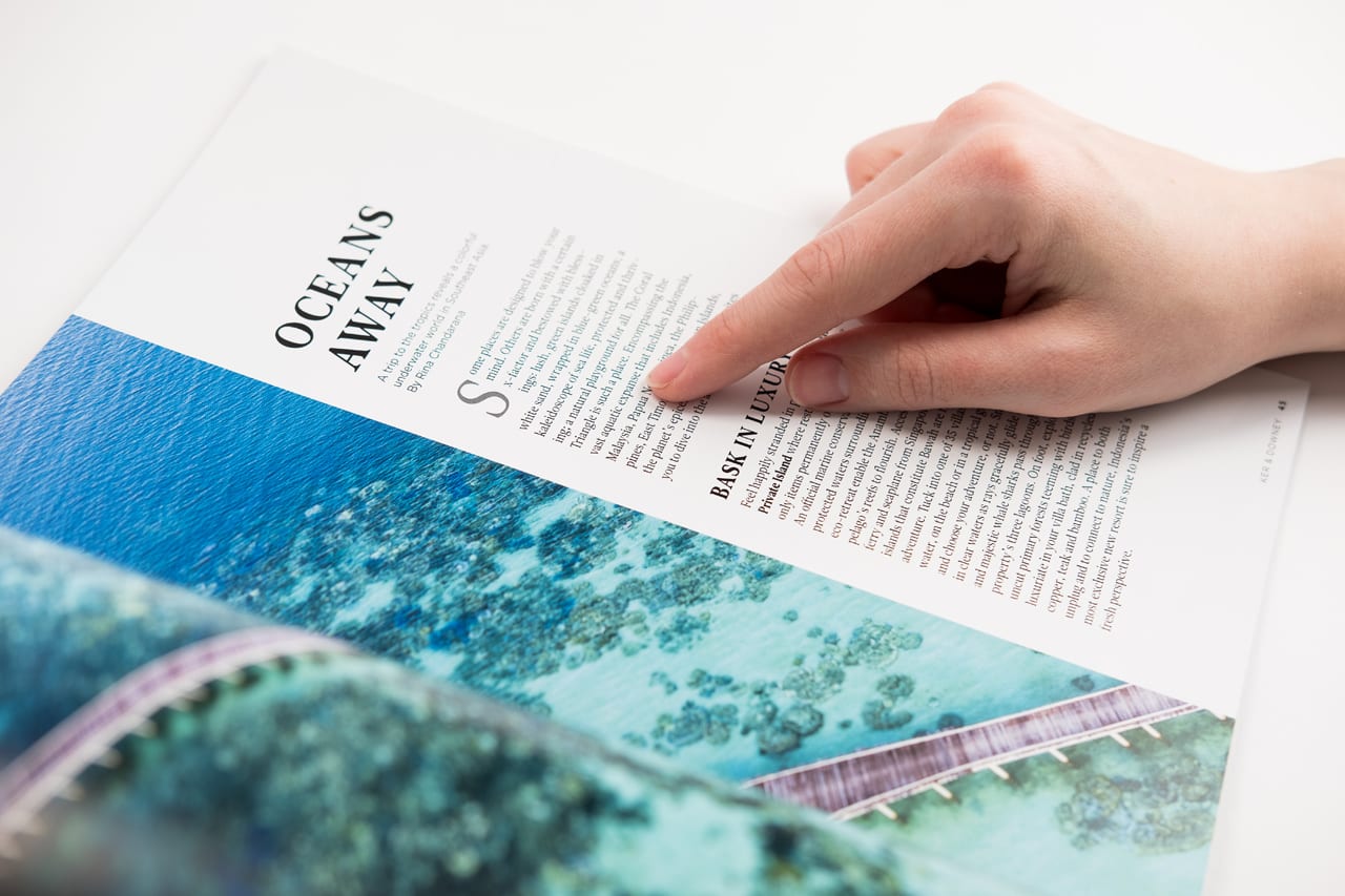 A travelog booklet laying open to ocean imagery and content with a hand pointing to a paragraph on the right.