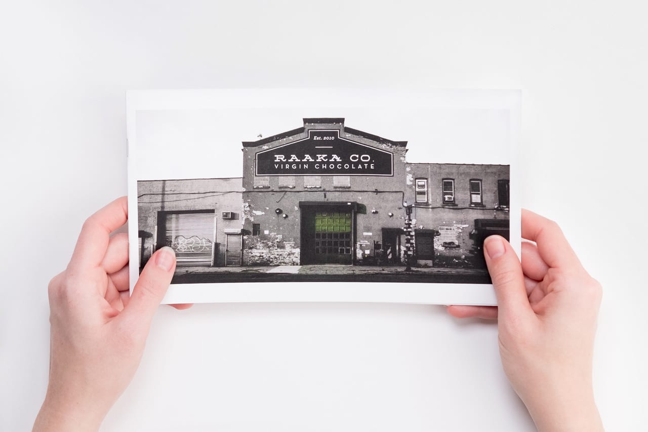 Two hands holding a marketing booklet printed with an image of a building with Raaka Co. Virgin Chocolate on it.