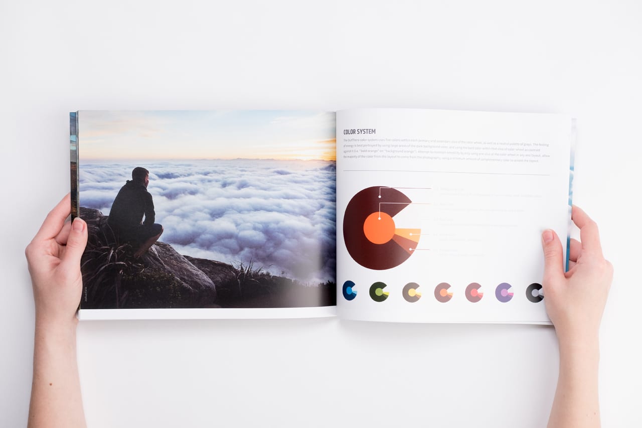 Two hands holding open a brand manual printed with a man on a cliff overlooking clouds and information about color.