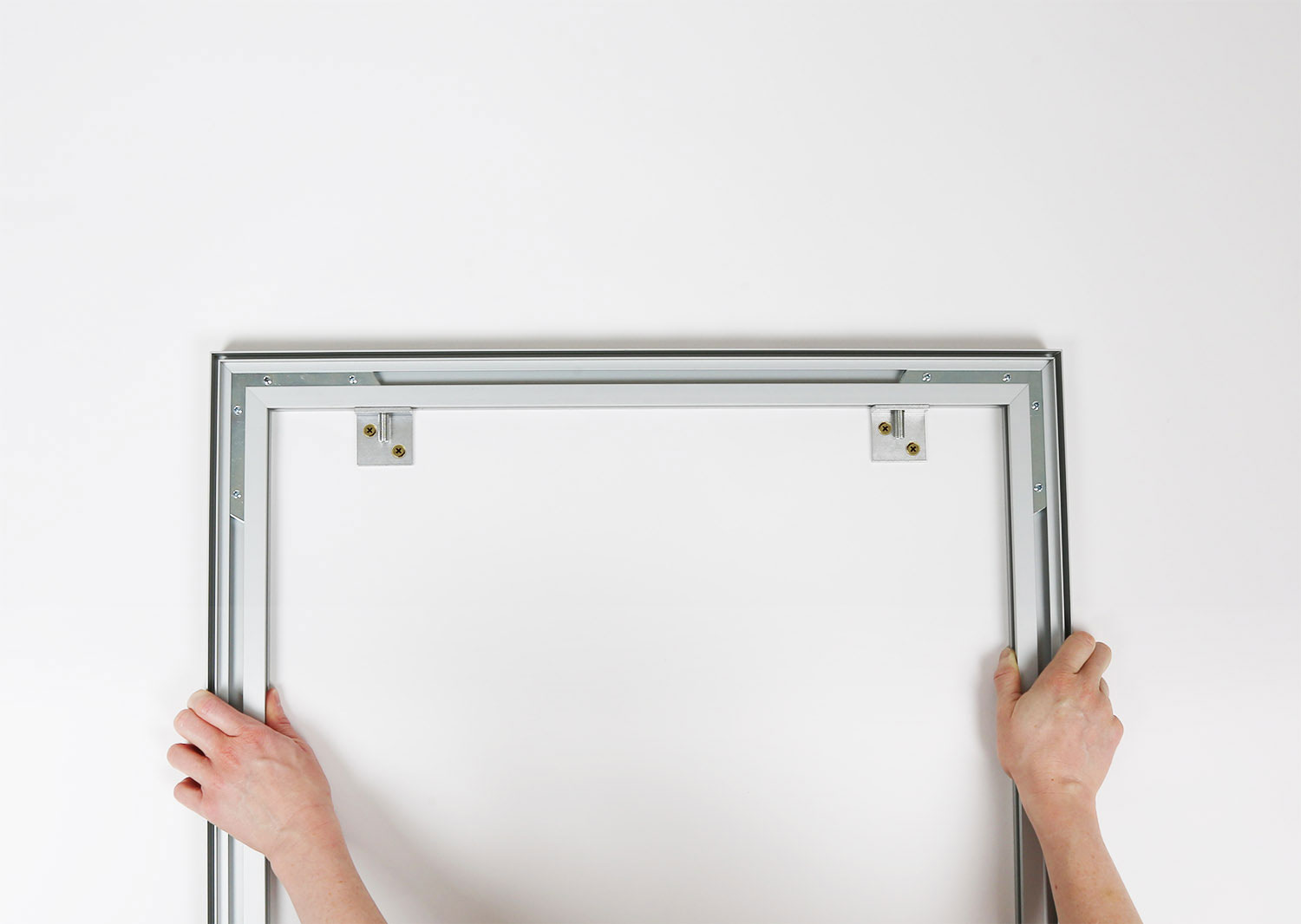 Two hands holding an aluminum frame for an SEG against a wall with its mounting hardware.