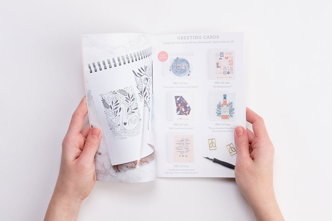 Two hands holding open a product catalog with images of and details for greeting cards.