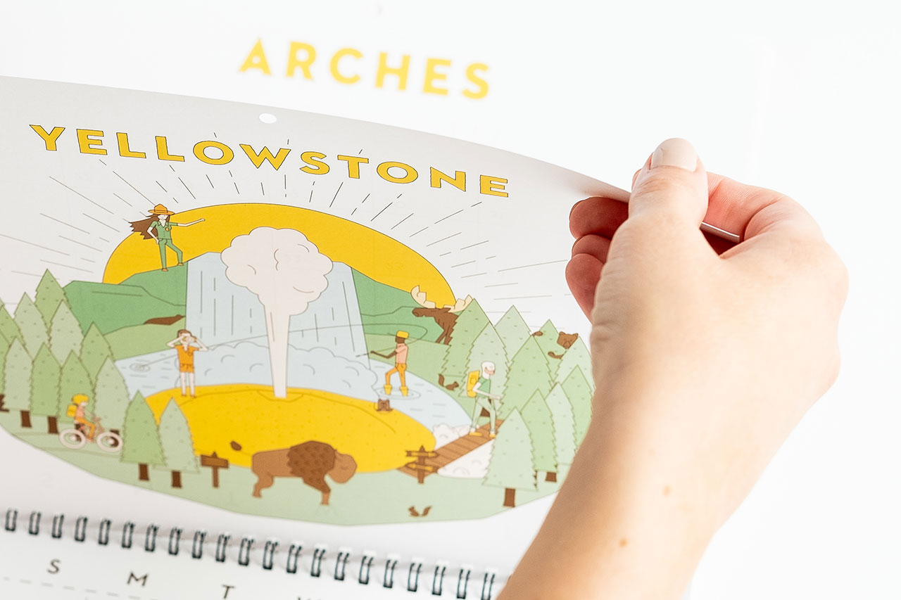 A hand turning the page of a custom calendar printed with national parks illustrations.