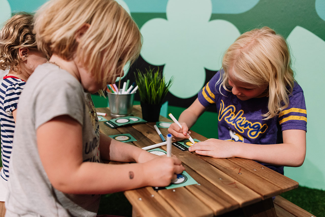 Three kids coloring at a wooden table with a green floral design on the wall behind them.