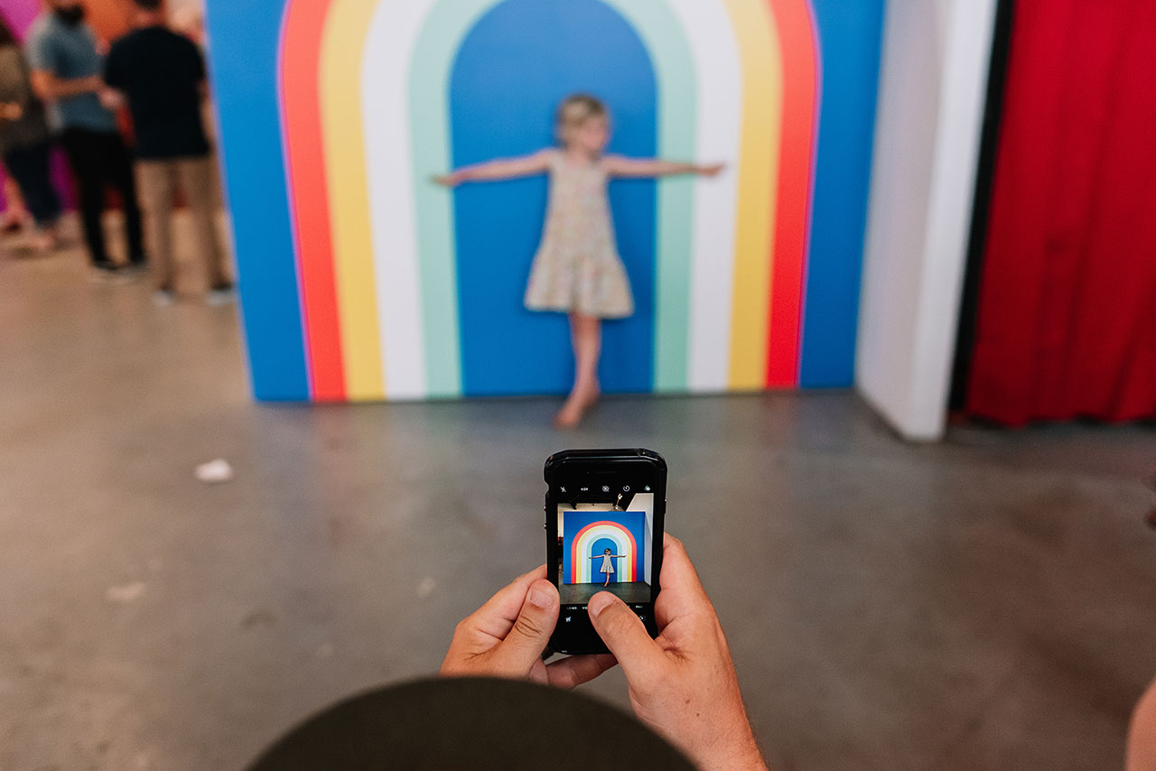 A little girl posing in front of a wall with a rainbow decal and a person taking a picture of her on their phone.