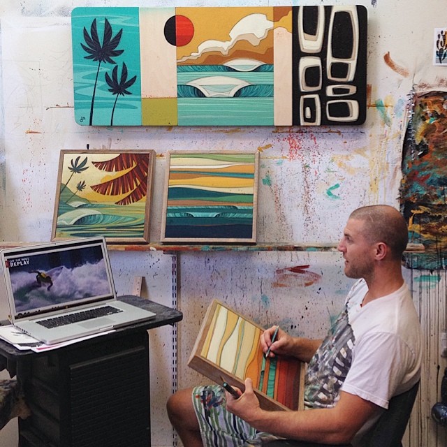 A man painting custom artwork next to a laptop on a table and more artwork hanging on the wall.