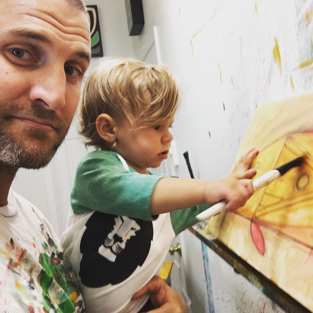 A man in an artist studio holding a baby with a paintbrush painting yellow color on a wall.