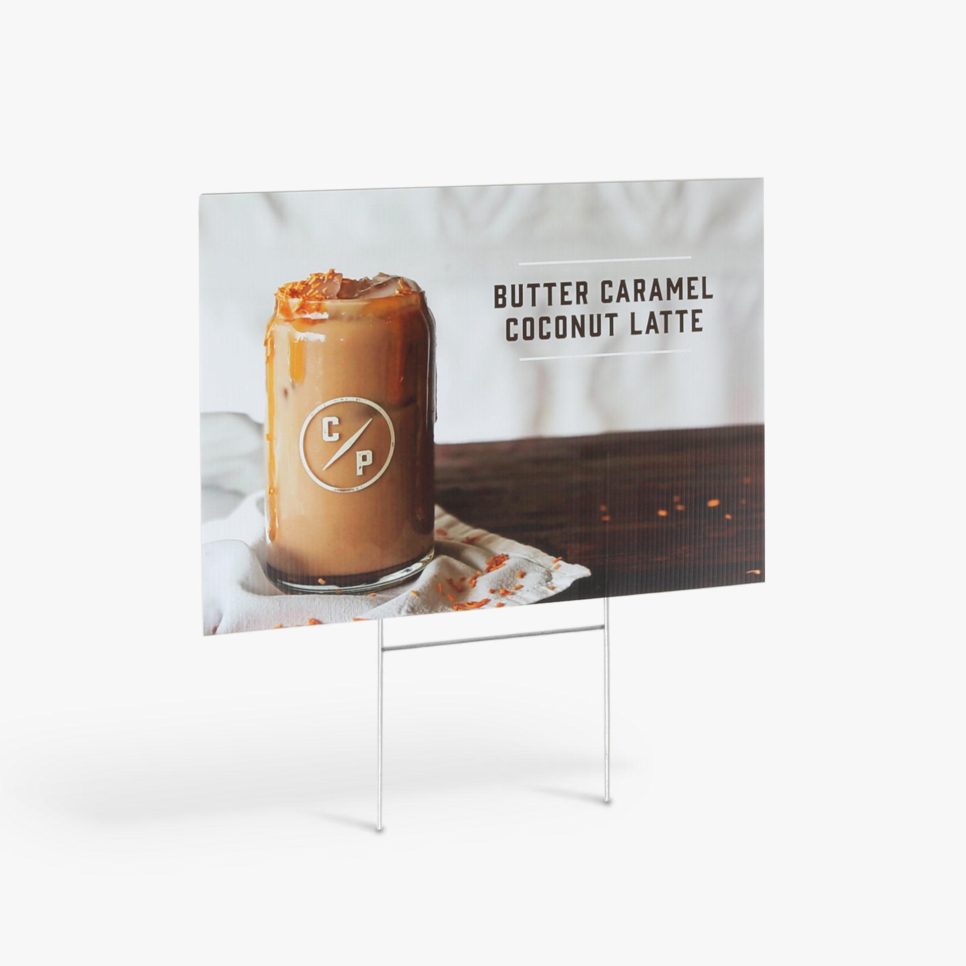A custom lawn sign printed with Butter Caramel Coconut Latte in brown next to an iced coffee.