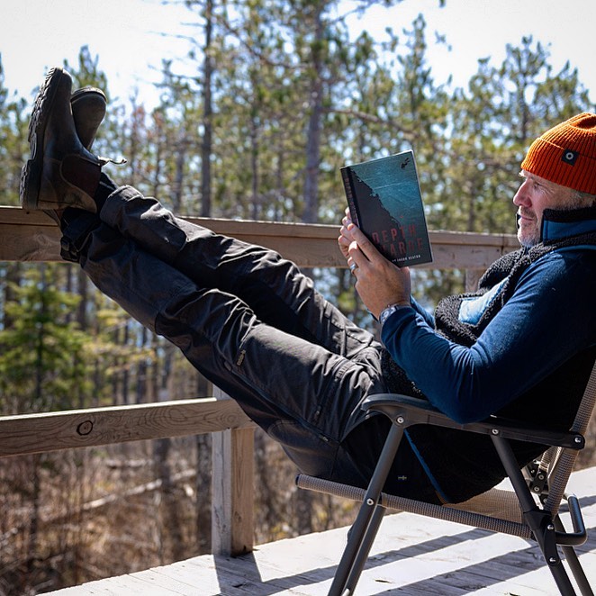 A man sitting in a chair reading a book on a porch overlooking a forest of trees.