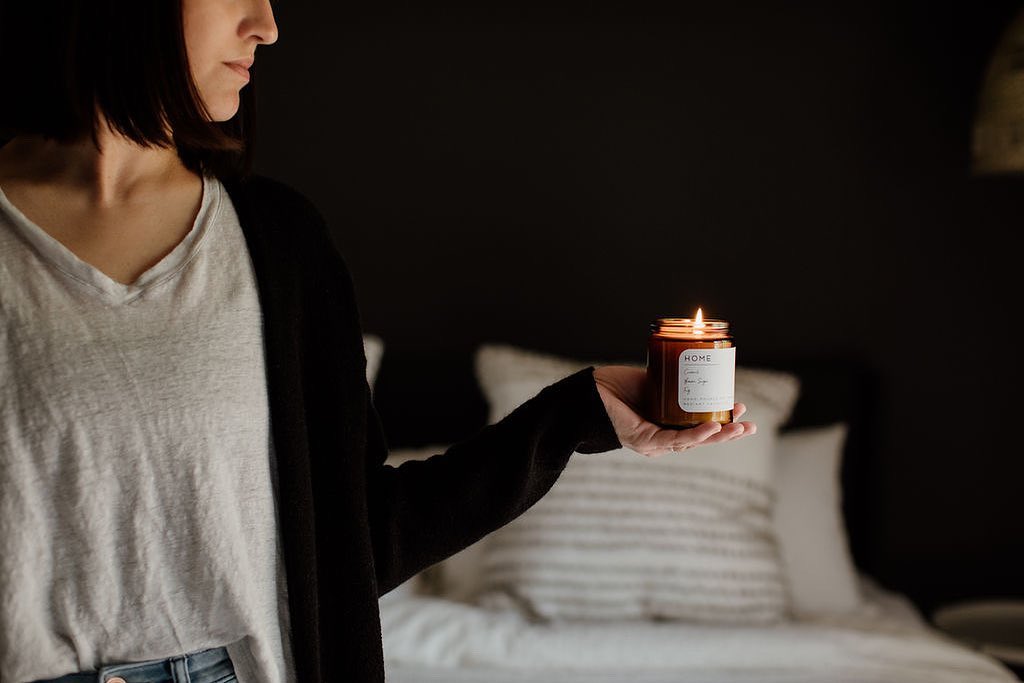 A woman in a white shirt and black cardigan holding a burning candle in a jar with a custom printed label.