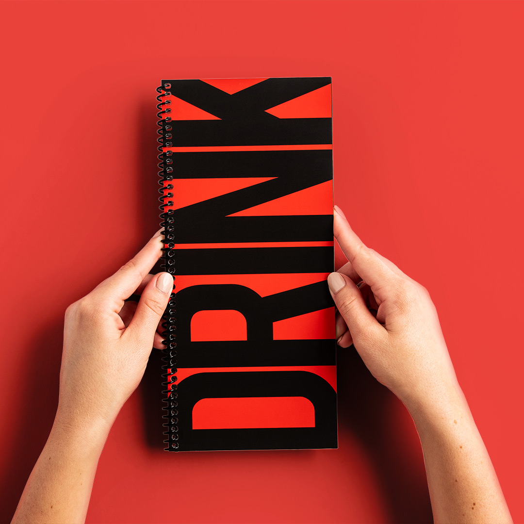 Two hands holding a custom drink menu with a red and black design and a spiral binding.