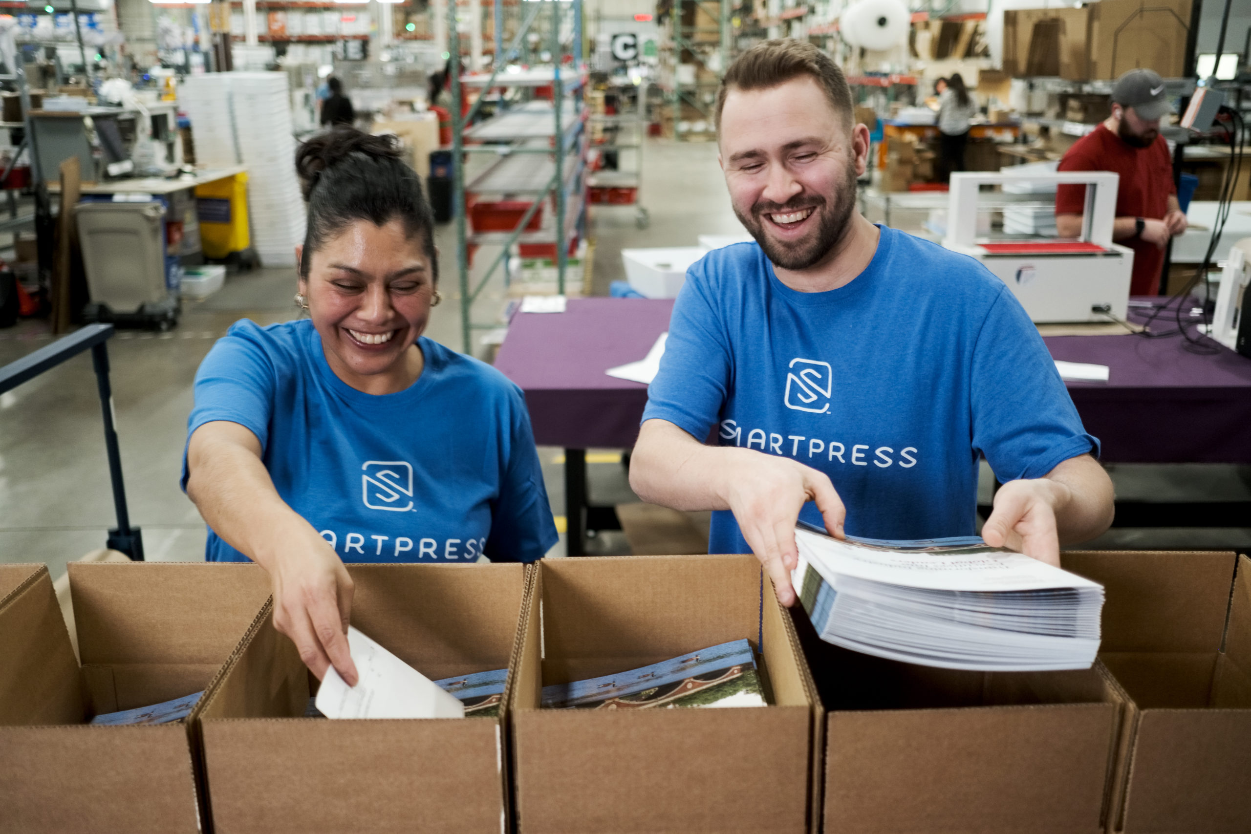 Two people wearing Smartpress T-shirts packing print products in cardboard boxes in a production facility.