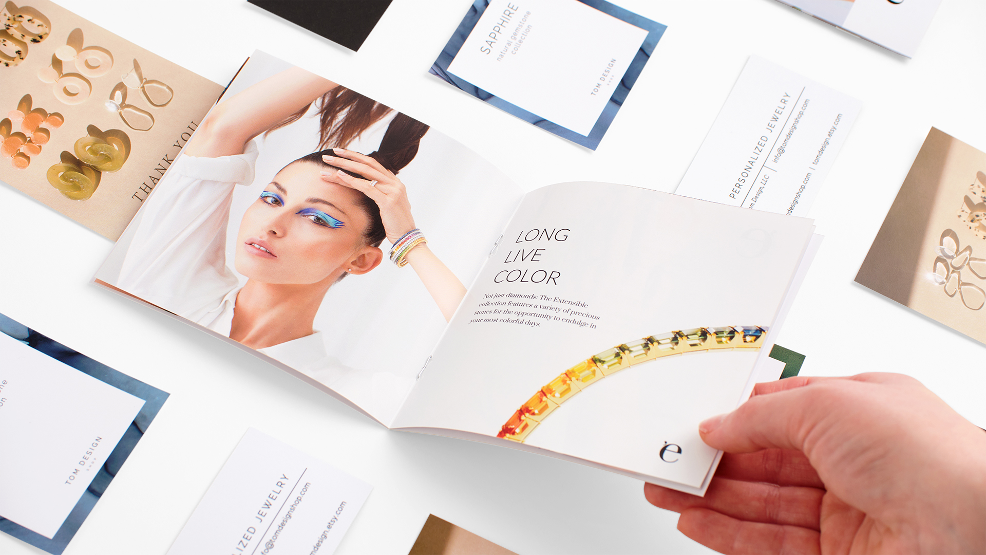 A hand holding open a jewelry product booklet with other jewelry print assets around it.